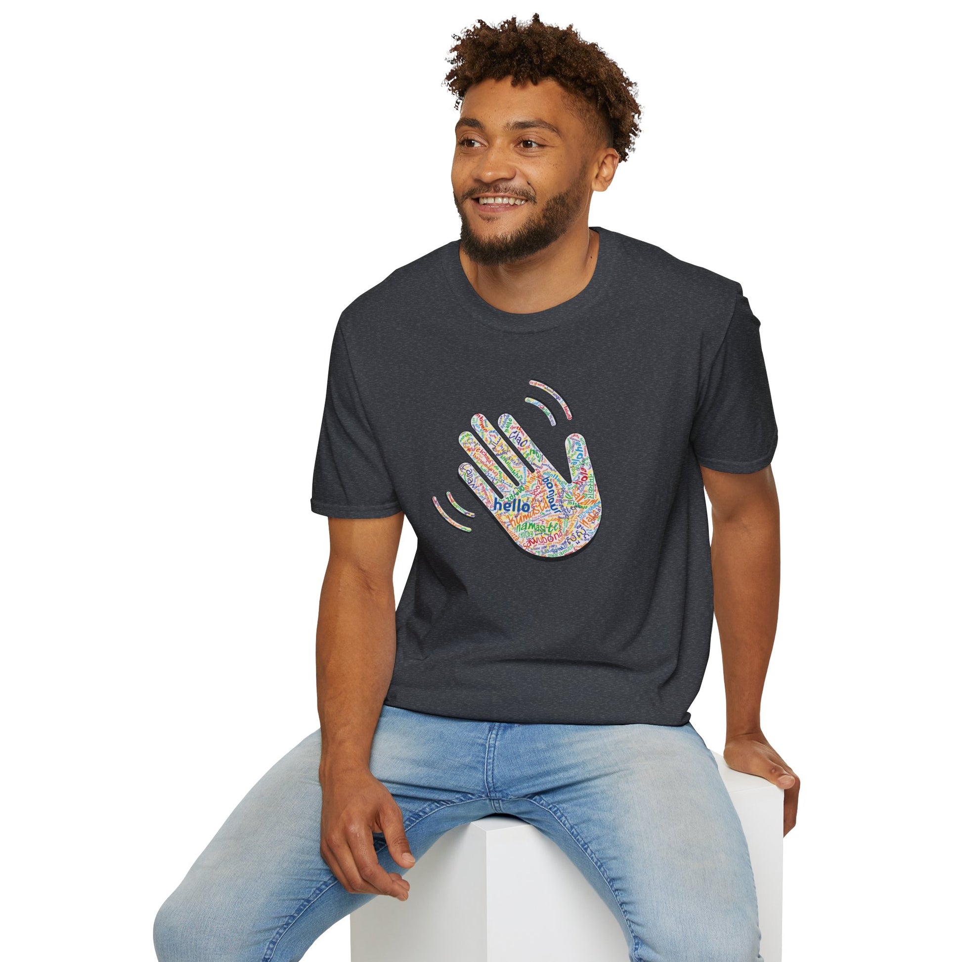 Man wearing a dark gray T-shirt with a colorful hand graphic filled with greetings from many languages, sitting on a white stool against a white background.