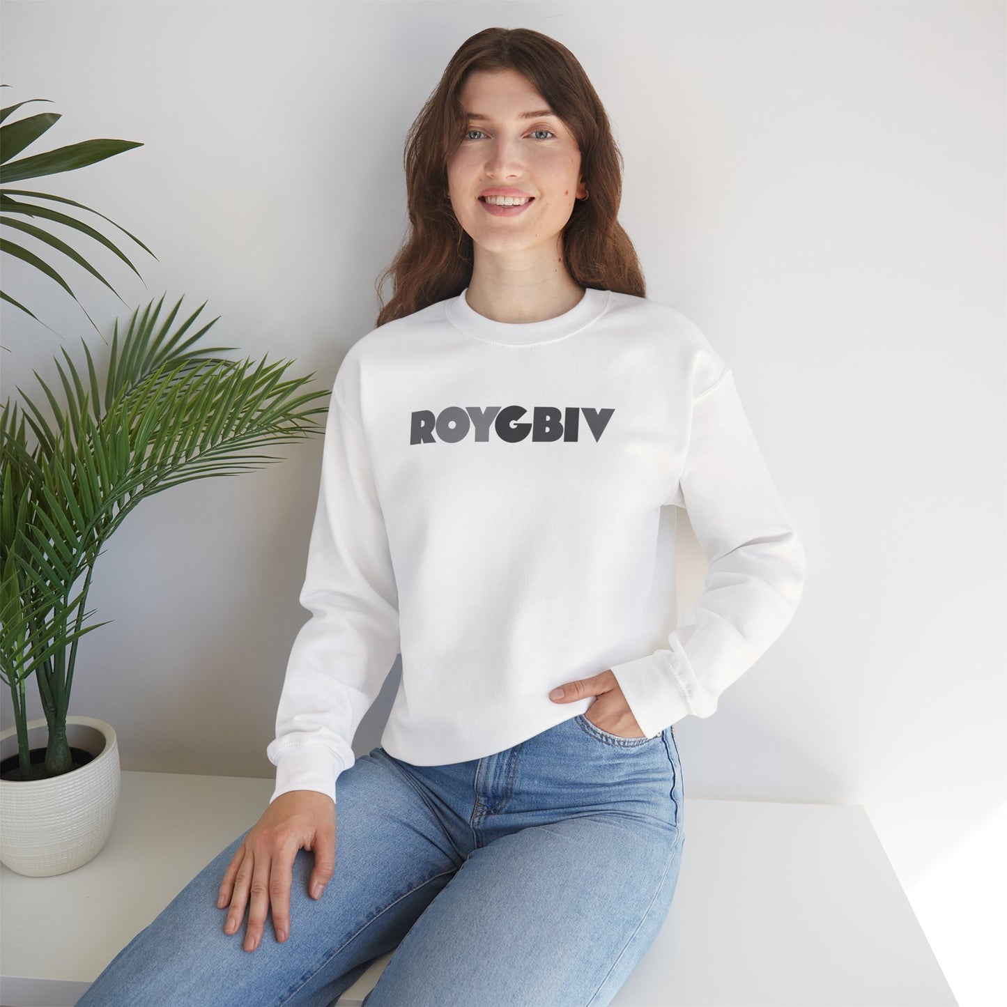 Woman wearing a white sweatshirt with 'ROYGBIV' text, sitting indoors with a plant in the background.