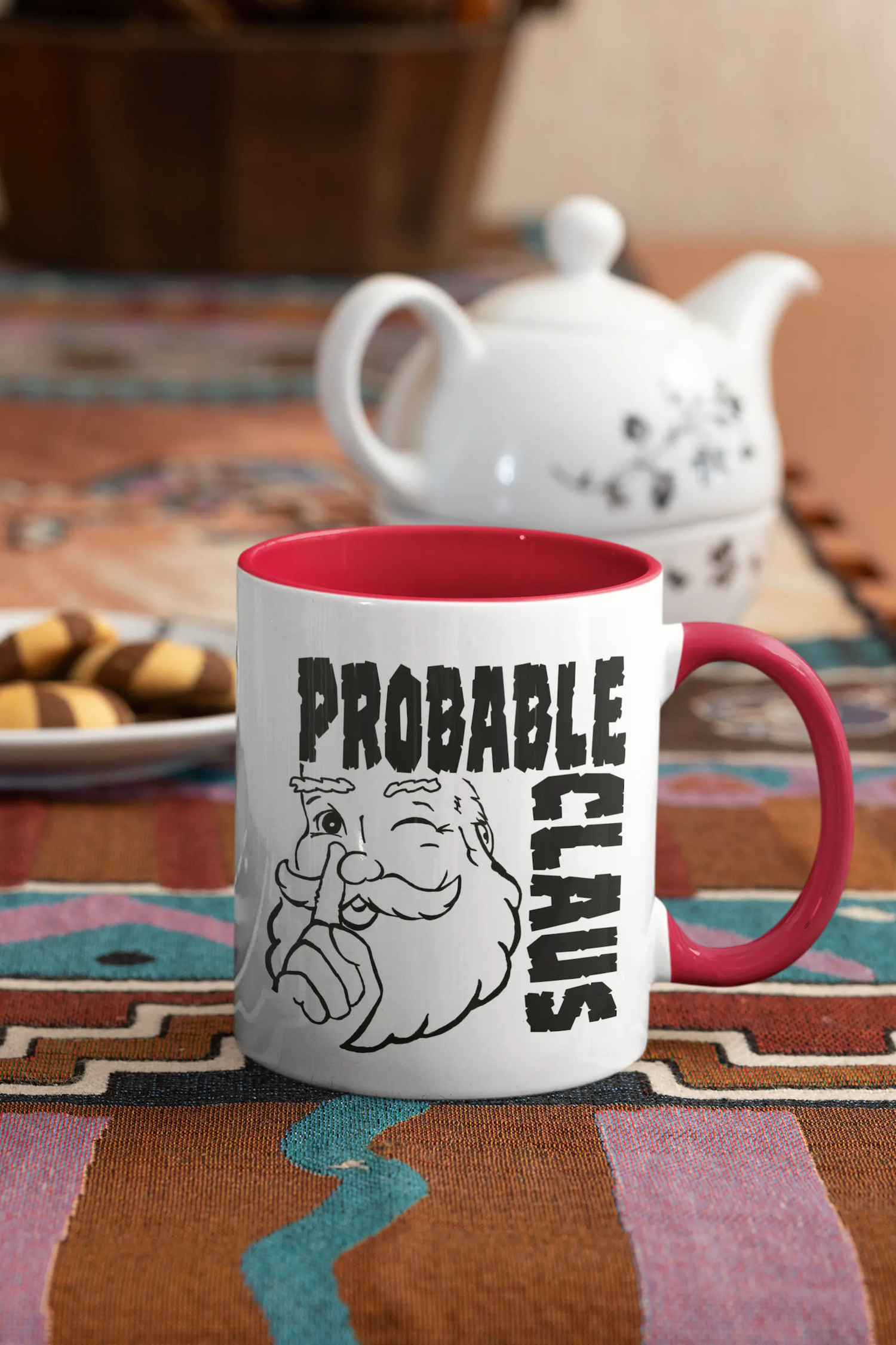 White mug with red handle and interior accent printed with Santa Claus giving a wink and the words 'Probably Claus' in large block letters.  Mug is on a cloth tablecloth and there is a teapot and a plate of cookies in the background.