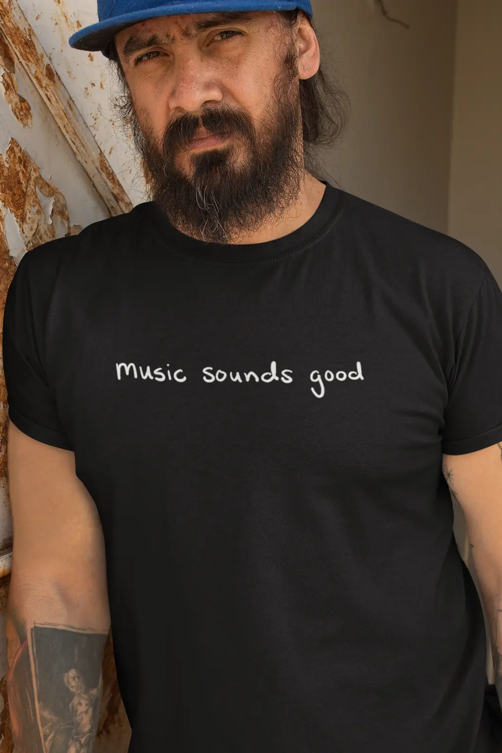Man wearing a black t-shirt with 'Music Sounds Good' text, leaning against a wall.