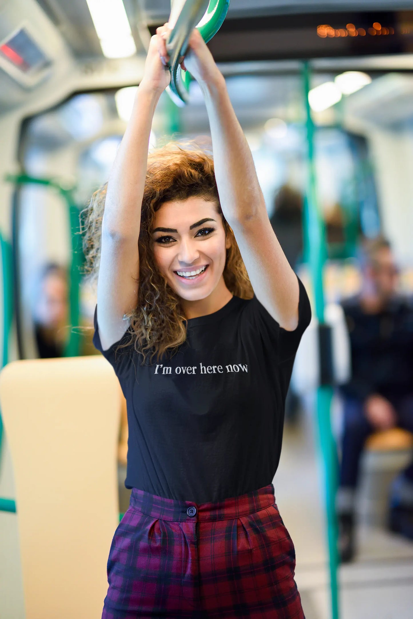 Woman on a train wearing a black "I'm Over Here Now" T-shirt, holding onto a handrail.