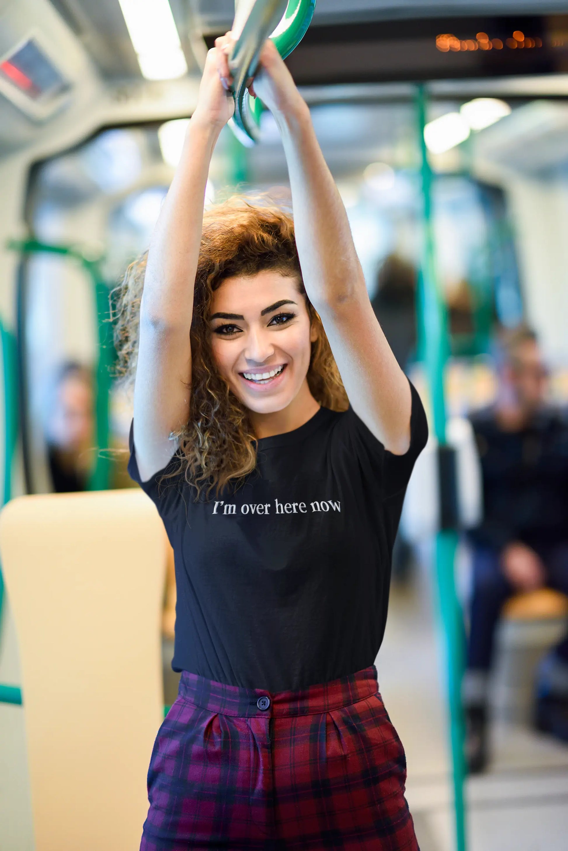 Woman on a train wearing a black "I'm Over Here Now" T-shirt, holding onto a handrail.