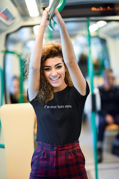Woman on a train wearing a black "I'm Over Here Now" T-shirt, holding onto a handrail.