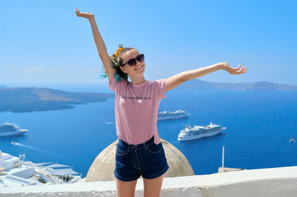 Person with arms outstretched in front of a scenic view with boats and a blue sky, wearing pink "I'm Over Here Now" tee shirt 
