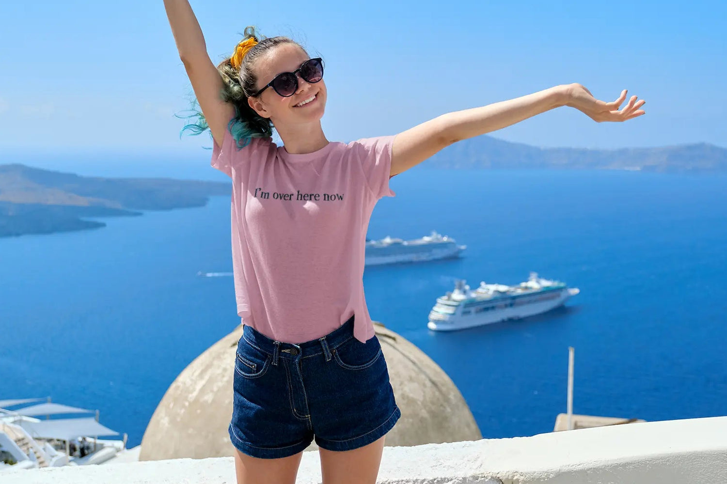 Person with arms outstretched in front of a scenic view with boats and a blue sky wearing a pink Think a Thunk Unisex Premium I'm Over Here Now Graphic T-shirt.