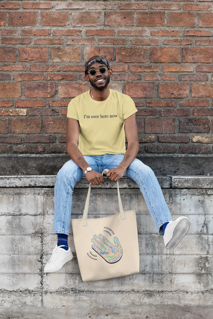Man wearing yellow "I'm Over Here Now" T-shirt, sitting on a wall with a tote bag featuring a handprint design.