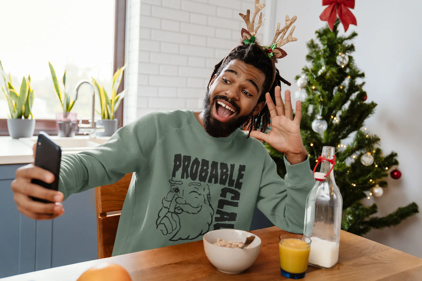 Man taking a selfie in a festive setting with a Christmas tree in the background, wearing green 'Probable Claus' sweatshirt.
