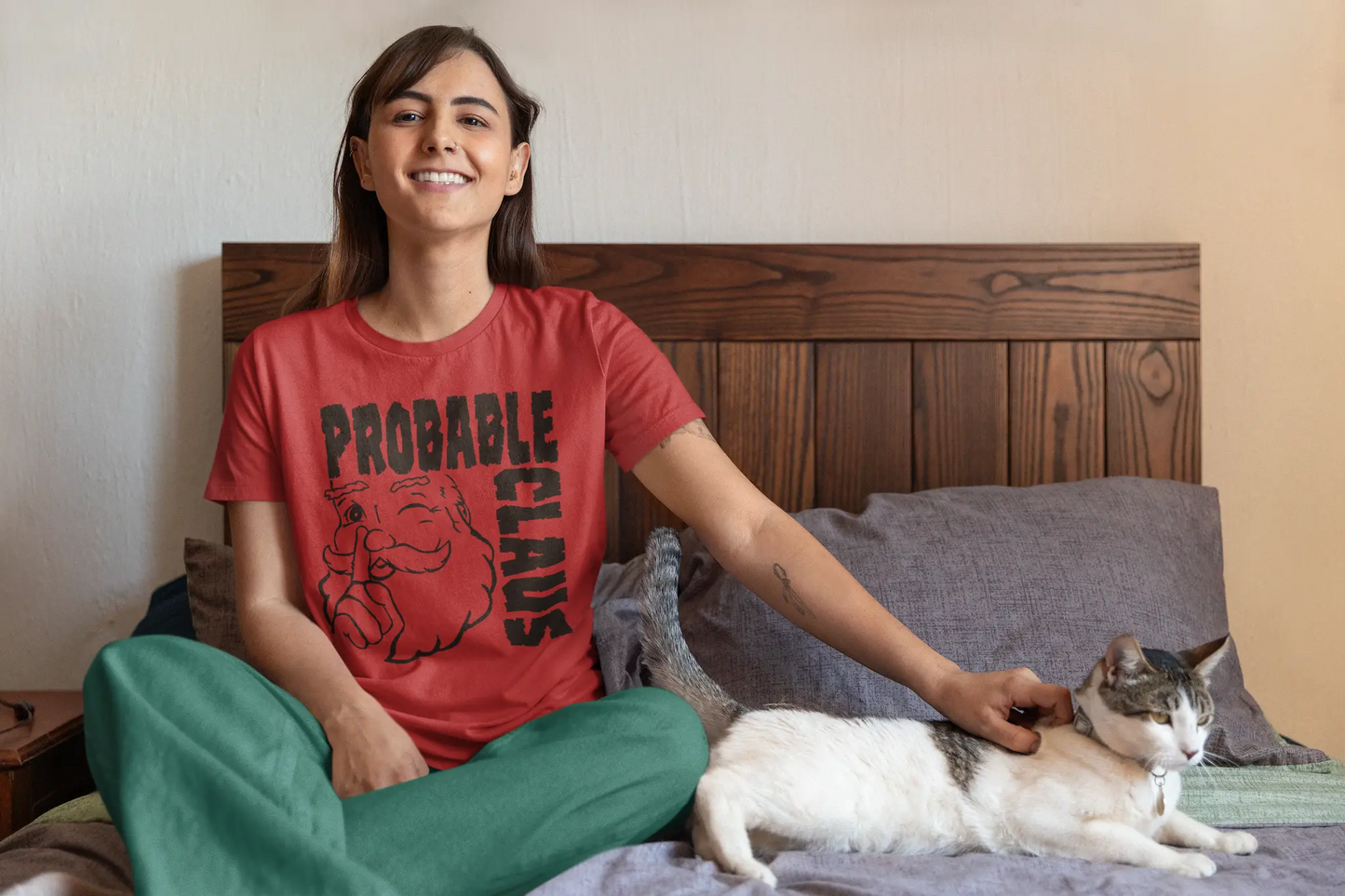 Woman sitting on a bed with a cat, wearing a red t-shirt with 'Probable Claus' graphic and green pants.