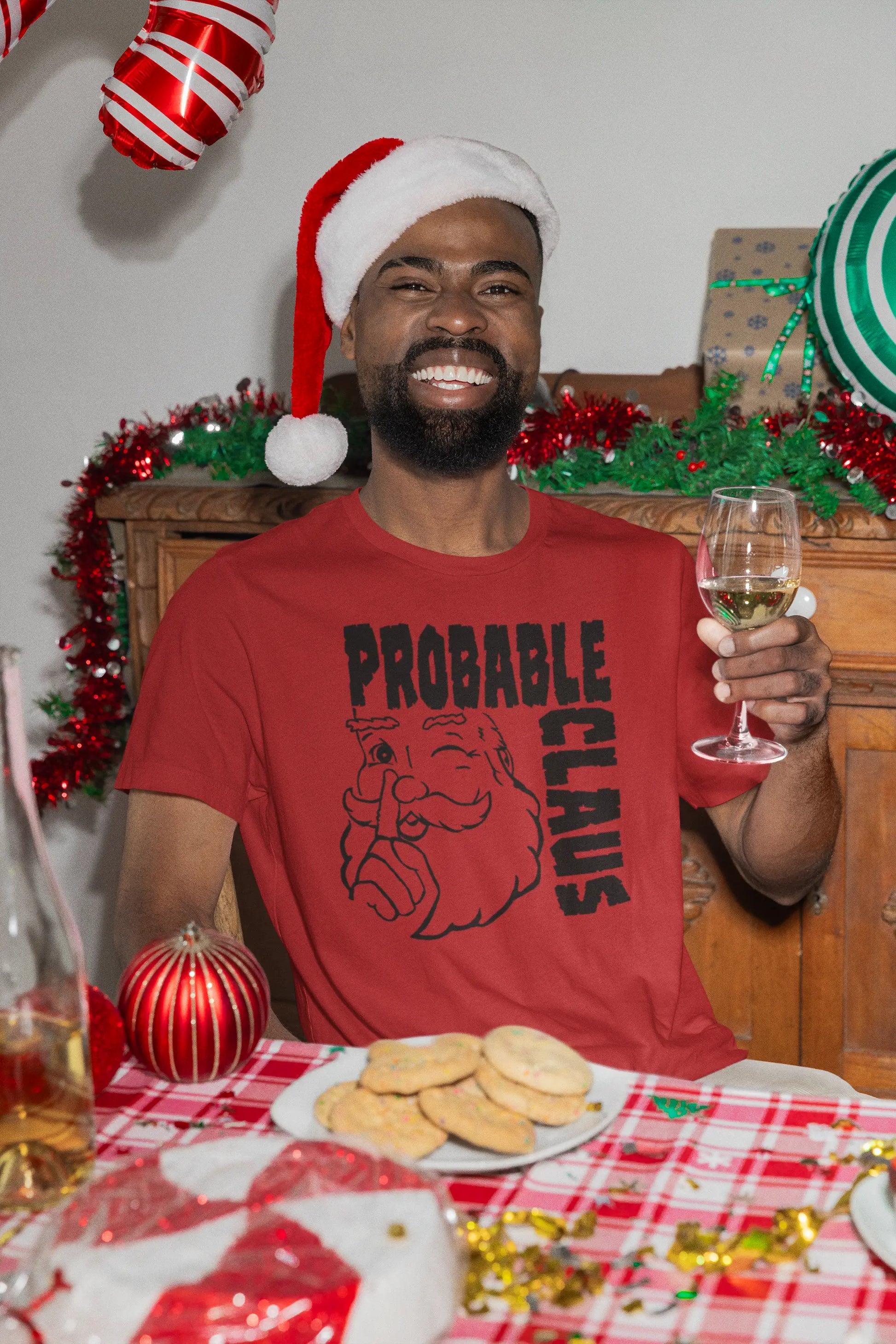Man wearing a Santa hat and 'Probable Claus' shirt holding a glass of wine at a Christmas-themed table.