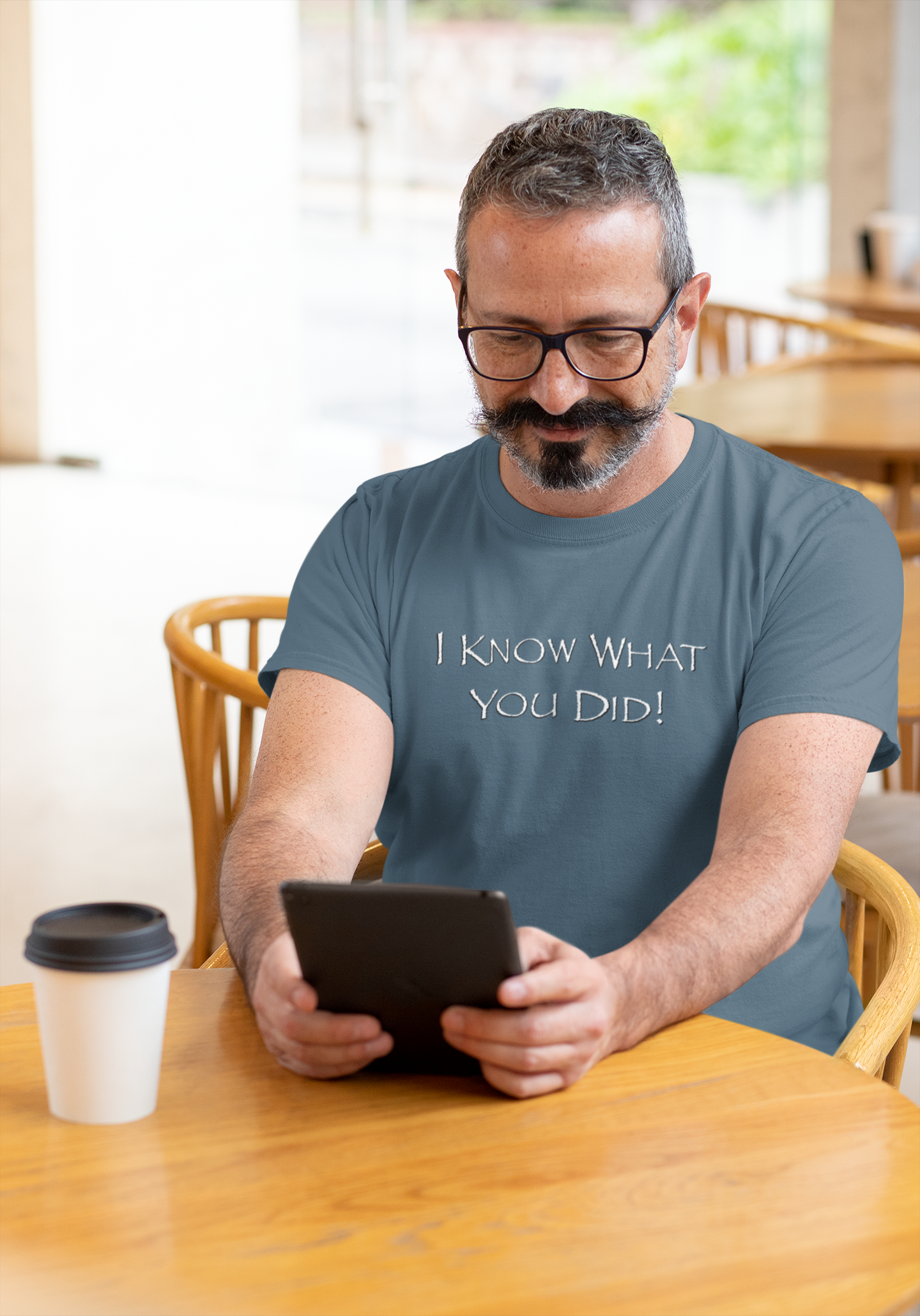 Man wearing a t-shirt with text, sitting at a table with a tablet and coffee.