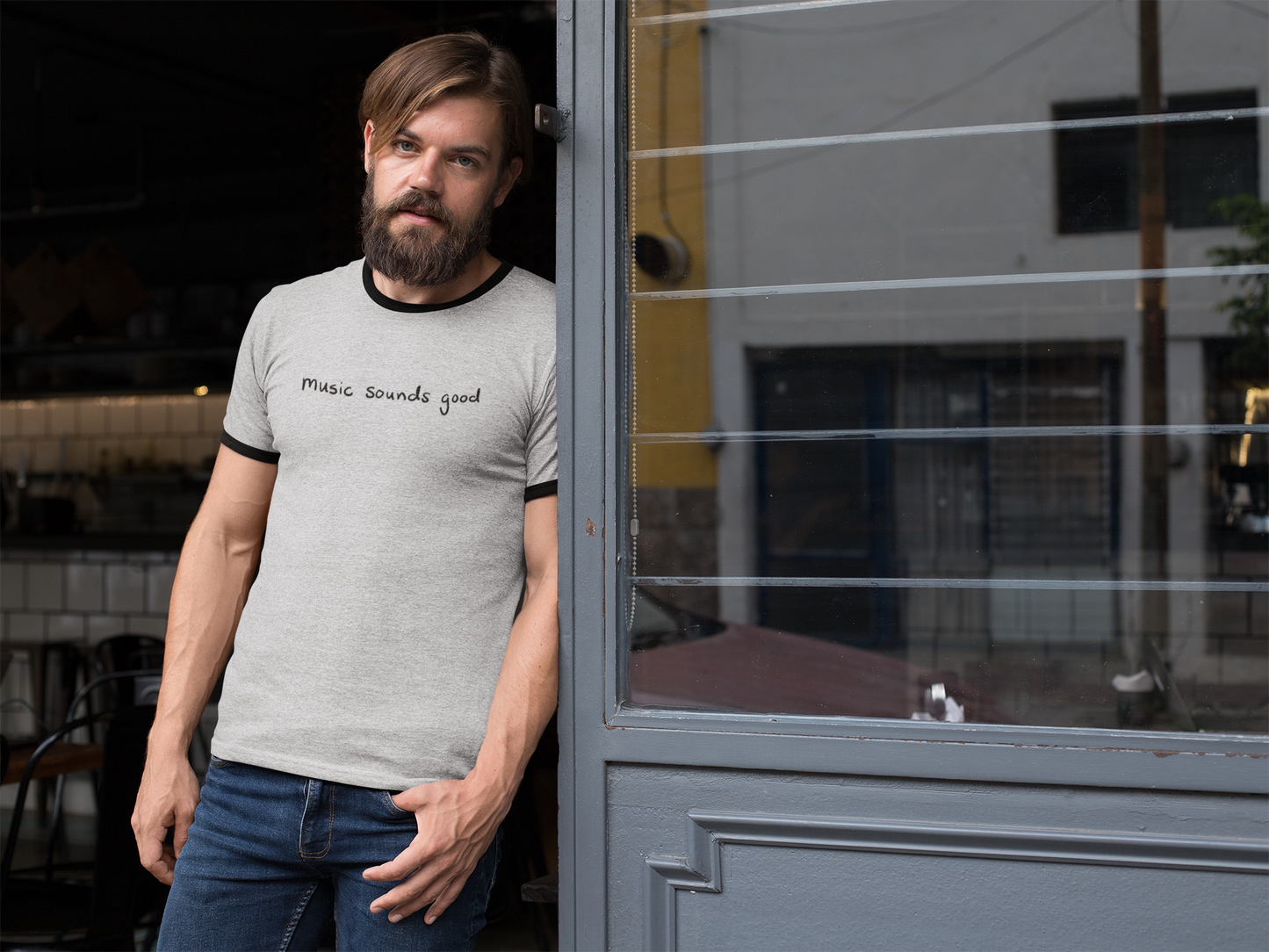 Man wearing a gray ringer t-shirt with the text 'music souunds good', standing next to a door.