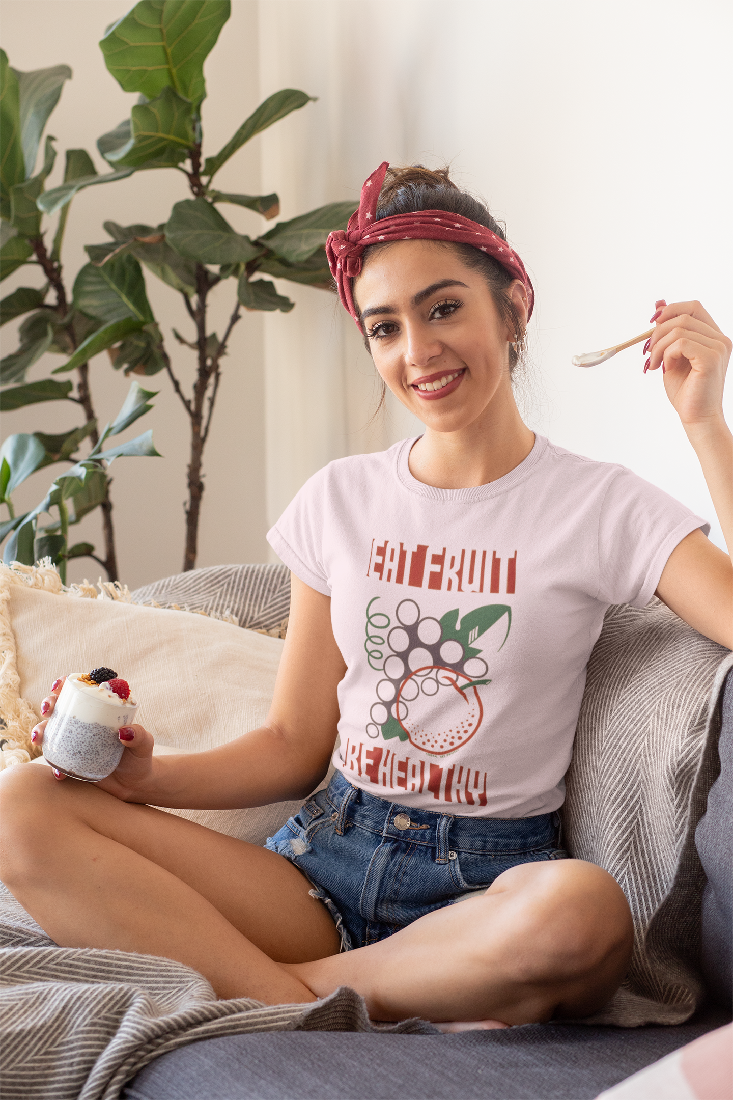 Woman sitting on a couch holding a cup and a spoon, wearing a t-shirt with a graphic design.