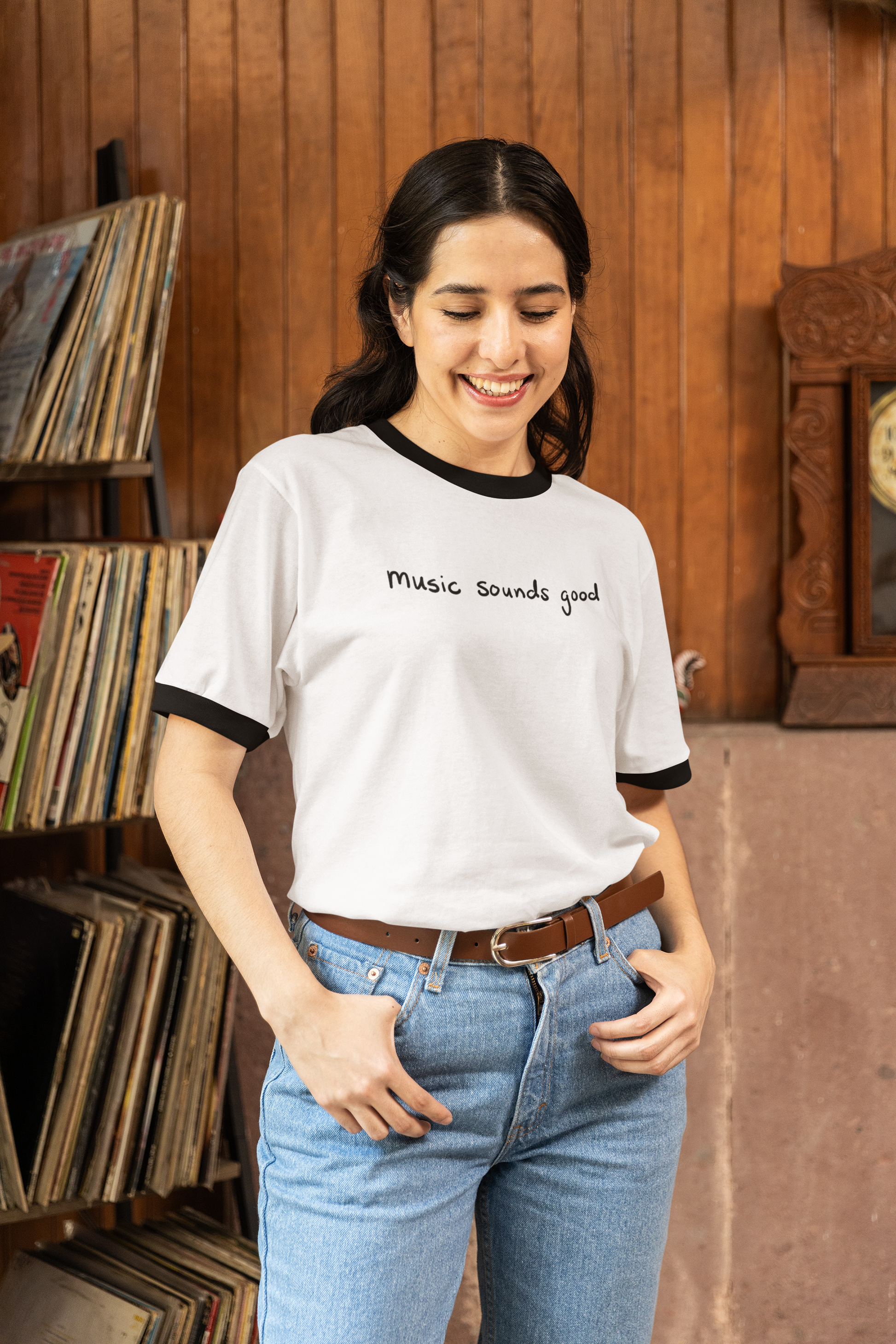 Woman wearing a white ringer t-shirt with 'music sounds good' text in front of shelves filled with records.