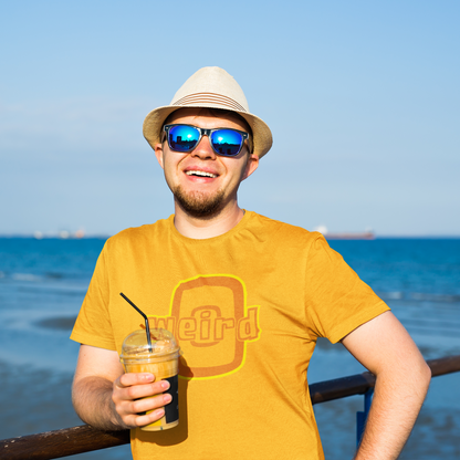 Man wearing a yellow t-shirt with a logo, sunglasses, and a hat, holding a drink by the ocean.