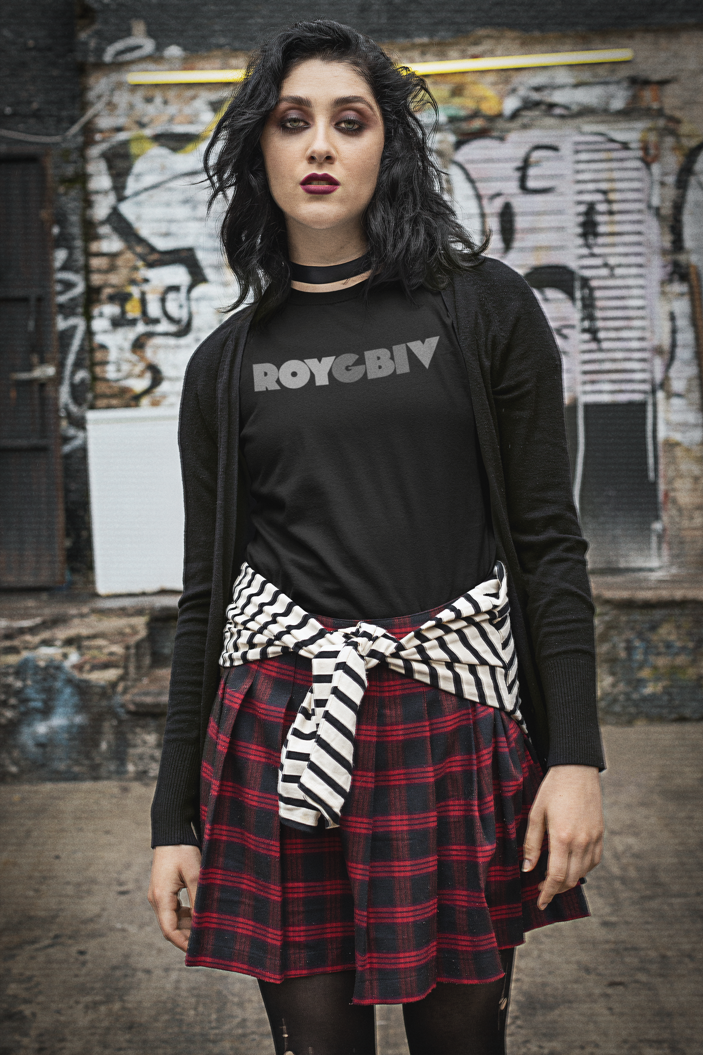 Person wearing a black 'ROYGBIV' shirt with a plaid skirt and zebra-striped sash in front of a graffiti-covered wall.