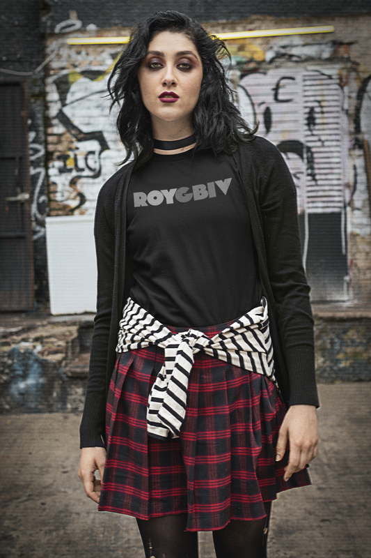 Person wearing a black 'ROYGBIV' shirt with a plaid skirt and zebra-striped sash in front of a graffiti-covered wall.