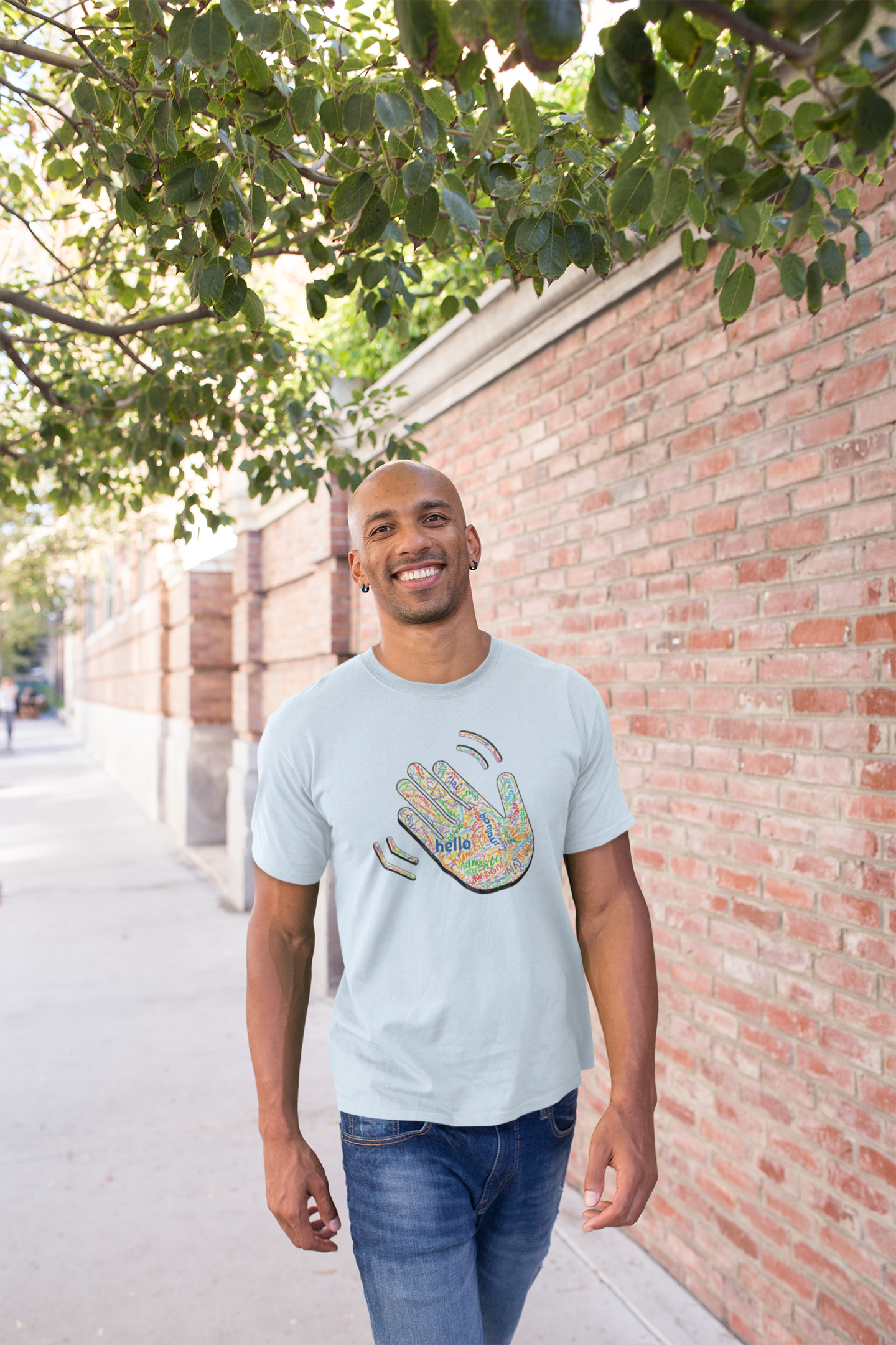 Man wearing a light blue T-shirt with a colorful hand graphic filled with greetings from many languages, standing outdoors near a brick wall and tree.