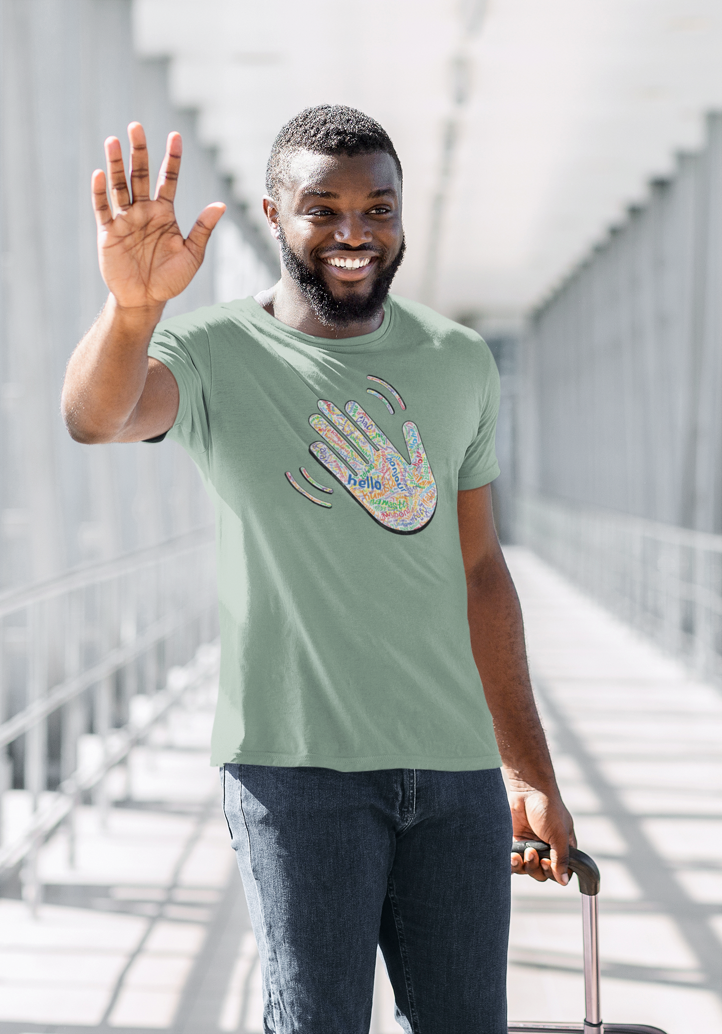 Man waving and smiling in a green T-shirt with a colorful hand graphic filled with greetings from many languages, standing on a bridge.