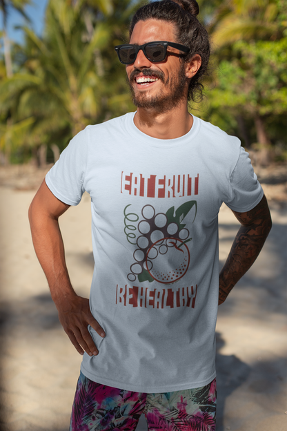 Man wearing a light blue t-shirt with a graphic design and the text "EAT FRUIT BE HEALTHY", standing on a beach.
