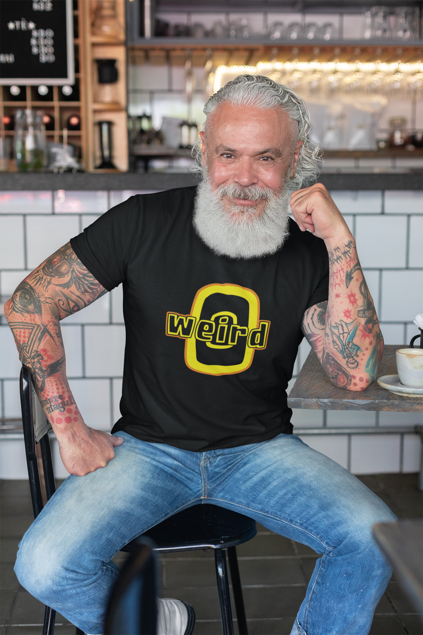 Man with tattoos wearing a black t-shirt with 'weird' printed on it, sitting in a cafe.
