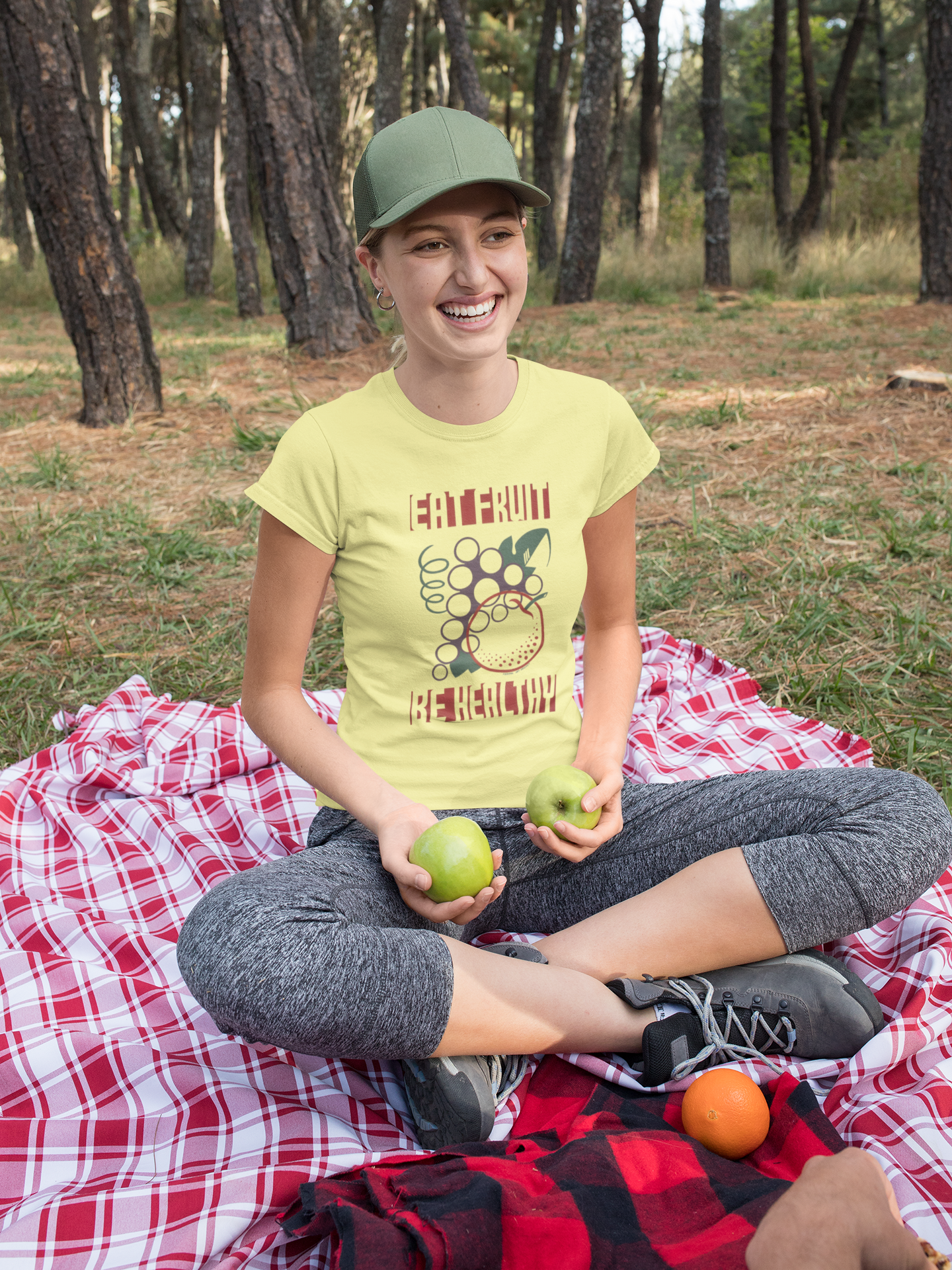 Person sitting on a red and white checkered blanket in a forest, holding apples and an orange.
