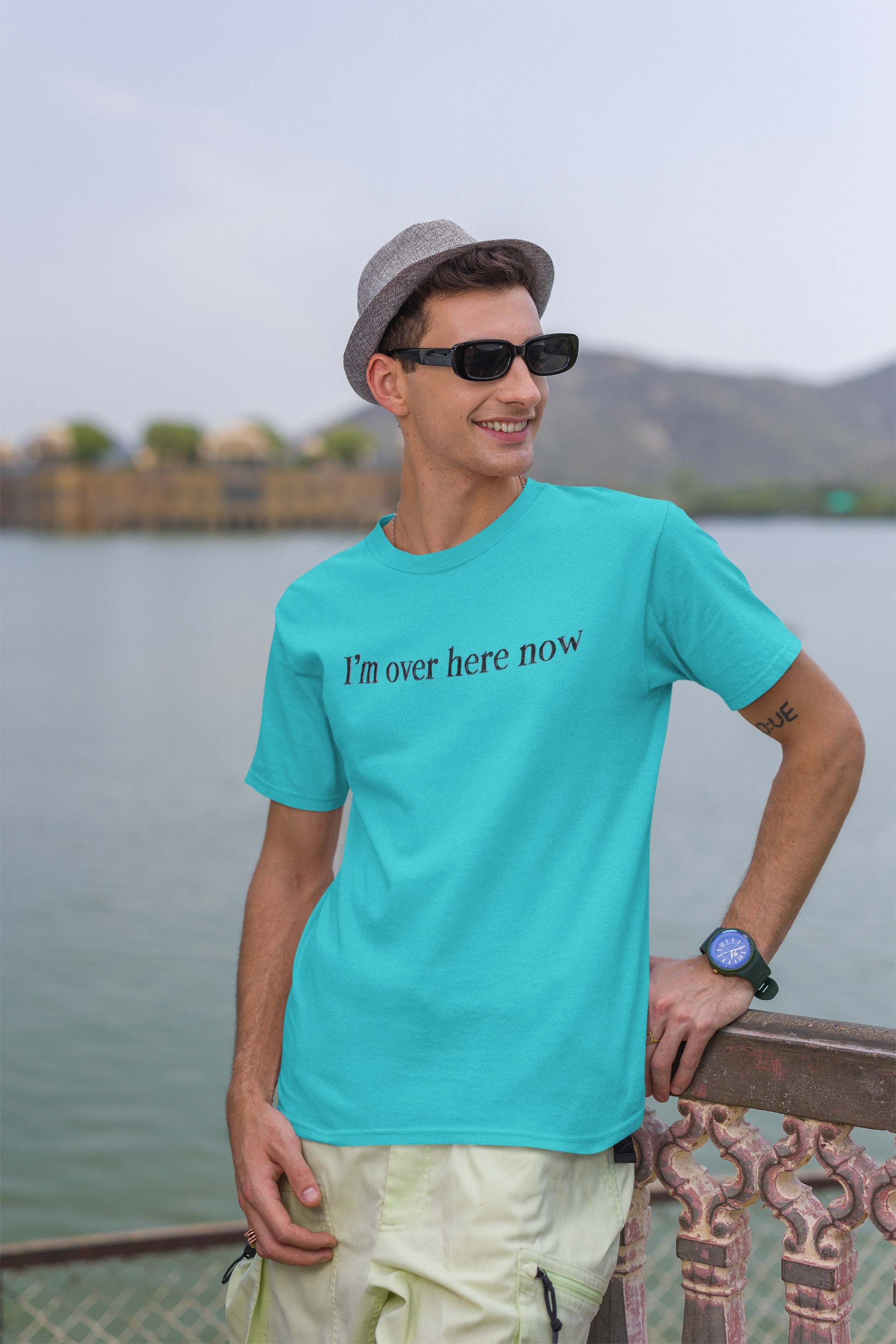 Man wearing a turquoise t-shirt with 'I'm over here now' text, sunglasses, and a hat, standing by a body of water.