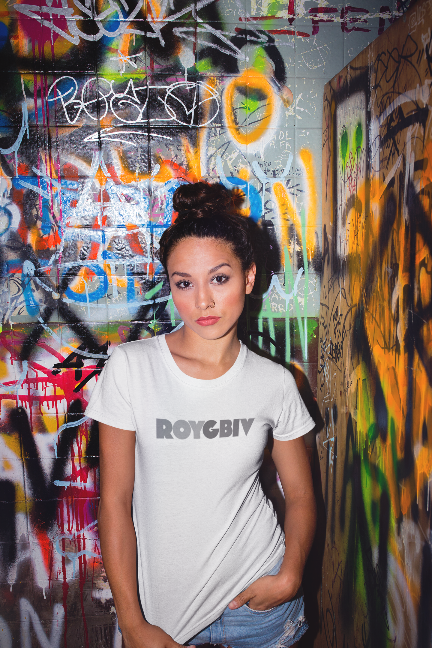 Woman wearing a white t-shirt with 'ROYGBIV' text in front of a graffiti-covered wall.
